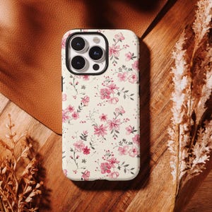 May include: A phone case with a floral design. The case has a cream background with pink flowers and green leaves. It has a black border and is on a wooden surface. Dried plants are also visible.