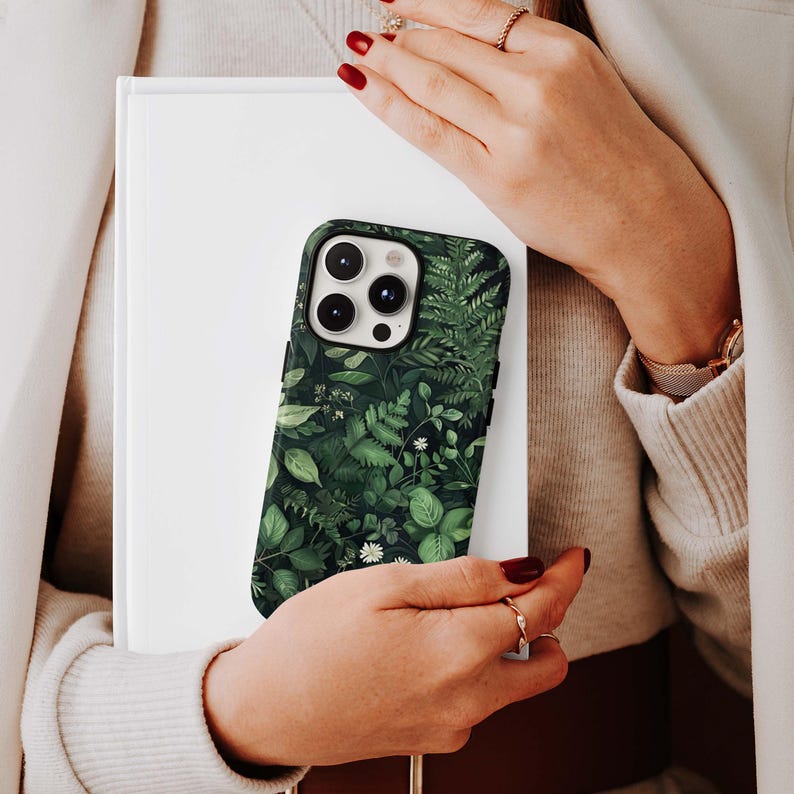 Puede incluir: Una funda de tel&eacute;fono con un dise&ntilde;o bot&aacute;nico verde oscuro con varias hojas y peque&ntilde;as flores blancas. La funda se sostiene contra un libro blanco, mostrando el intrincado patr&oacute;n de plantas.