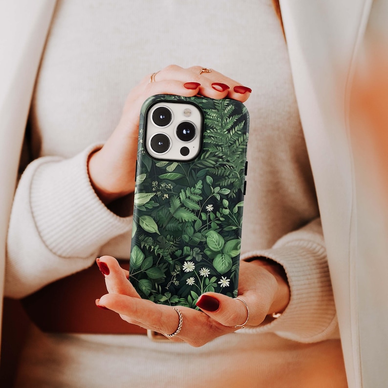Puede incluir: Funda de tel&eacute;fono verde oscuro con un dise&ntilde;o bot&aacute;nico de hojas verdes y flores blancas. La funda tiene un acabado brillante y un recorte para la c&aacute;mara. La funda del tel&eacute;fono la sostiene una persona.