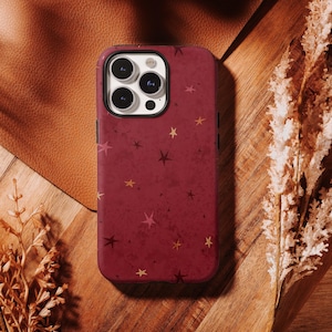 May include: A burgundy phone case with a star pattern. The case has a matte finish and features various sizes of gold and pink stars. The phone case is on a wooden surface with dried plants.