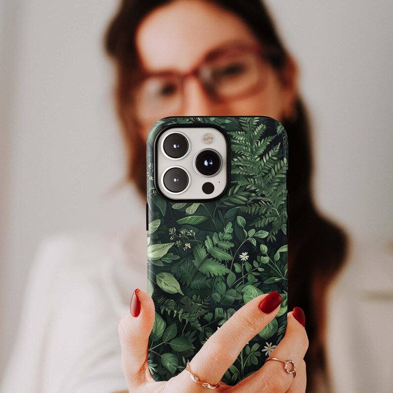 Puede incluir: Funda de tel&eacute;fono inteligente con un dise&ntilde;o bot&aacute;nico con varios tonos de hojas verdes y flores blancas. La funda tiene un borde negro y un recorte para la c&aacute;mara. La funda del tel&eacute;fono se sostiene frente a un fondo borroso.