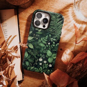 Puede incluir: Funda de tel&eacute;fono inteligente con un dise&ntilde;o bot&aacute;nico verde oscuro que presenta varias hojas y peque&ntilde;as flores blancas. La funda tiene un borde negro y un recorte para la c&aacute;mara. El dise&ntilde;o es detallado y realista.