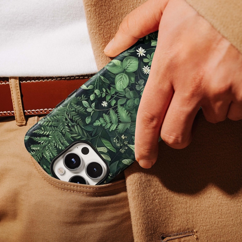 Puede incluir: Una funda de tel&eacute;fono inteligente con un dise&ntilde;o bot&aacute;nico verde oscuro con varias hojas y flores blancas. La funda es parcialmente visible en un bolsillo, mostrando su intrincado patr&oacute;n y paleta de colores. La funda tiene un m&oacute;dulo de c&aacute;mara con tres lentes.