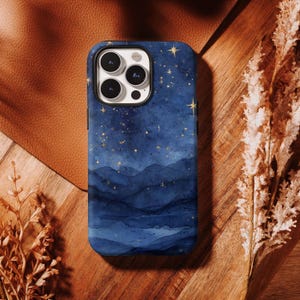 May include: A blue phone case with a watercolor design of a starry night sky. The case features a dark blue background with lighter blue mountain silhouettes and gold star accents. The case is on a wooden surface.