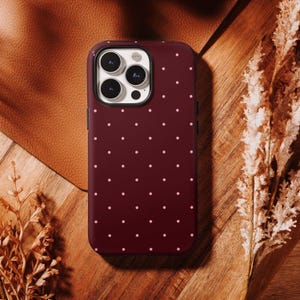 May include: A burgundy phone case featuring a repeating pattern of small, light pink polka dots. The case has a black border around the camera lenses and a black frame. The case is on a wooden surface.
