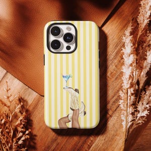 May include: A phone case featuring a vertical yellow and white striped pattern. The central graphic depicts a pair of legs wearing white cowboy boots, with one boot balancing a blue martini glass garnished with a lime slice on its toe.