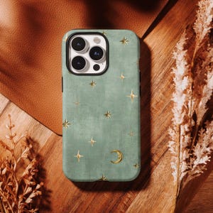 Peut inclure: Coque de téléphone vert sauge ornée d'étoiles dorées et d'un croissant de lune. Le boîtier a une bordure noire et est posé sur une surface en bois.