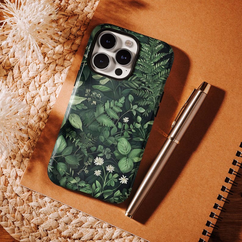 Puede incluir: Una funda de tel&eacute;fono con un dise&ntilde;o bot&aacute;nico verde vibrante que presenta varias hojas y peque&ntilde;as flores blancas. Un bol&iacute;grafo plateado descansa sobre un cuaderno marr&oacute;n junto a la funda del tel&eacute;fono. La funda tiene un acabado brillante.