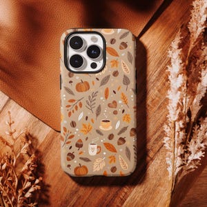 Peut inclure: Une coque de smartphone sur fond beige clair avec un motif répétitif de citrouilles orange, de glands marron, de feuilles d'automne aux tons jaunes, orange et marron, ainsi que de petites tasses blanches ornées de cœurs et de vapeur.
