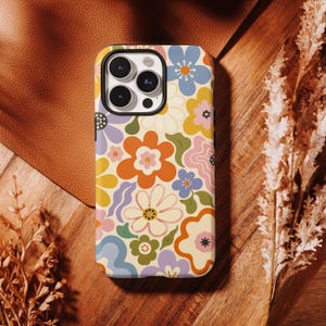 Pode incluir: Uma capa de telefone colorida com um padrão floral retrô. A capa apresenta uma variedade de flores em tons de laranja, amarelo, azul, rosa e roxo sobre um fundo creme. A capa é projetada para um smartphone.