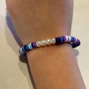 Könnte beinhalten: Ein Perlenarmband mit einer Mischung aus blauen, lila und weißen Polymer Clay Perlen. Das Armband ist mit drei weißen Perlen verziert. Das Armband hat einen Umfang von etwa 18 cm.