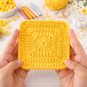 Op de afbeelding: Een vierkant, heldergeel gehaakt proeflapje wordt tussen twee handen gehouden. Het haakwerk heeft een textuurpatroon. Andere knutselspullen, waaronder knopen, garen en haaknaalden, zijn zichtbaar op de achtergrond.