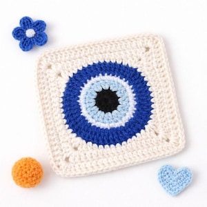 May include: Crocheted square with a blue eye design, surrounded by a cream-colored border. A blue flower, an orange ball, and a blue heart are scattered around the square. The handmade item is likely a decorative piece or part of a larger craft project.
