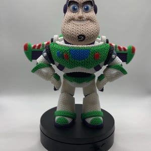 Puede incluir: Una figura de ganchillo de un personaje guardián espacial con capucha morada, peto verde y blanco y alas extendidas. La figura tiene ojos azules, un hoyuelo en la barbilla y se apoya sobre una base circular negra con un cable conectado.