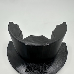 May include: A black 3D-printed object with the text "MP40" on the base. The object has a curved, semi-circular base and a U-shaped top section. The surface has visible layer lines from the printing process.