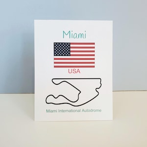 May include: White card featuring the word "Miami" in teal, the American flag, and the word "USA" in red. Below is a black outline of the Miami International Autodrome race track. The text "Miami International Autodrome" is at the bottom.