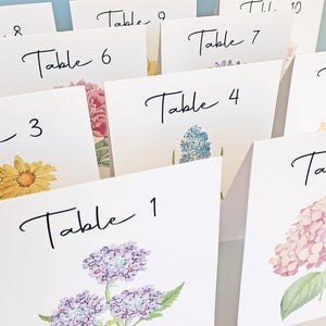 May include: White table cards with floral watercolor illustrations and the word "Table" in elegant black script. Each card is numbered, featuring different flowers like hydrangeas, daisies, and hyacinths, perfect for event decor.