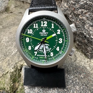 May include: A stainless steel wristwatch with a green face and black leather strap. The watch face displays white numerals, a crown emblem, and the text "ПОЛЕТ СИГНАЛ". The watch has two side buttons and a black display stand.