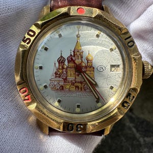 May include: A gold-toned wristwatch with a brown leather strap. The watch face features a detailed illustration of a cathedral with red and gold domes. The watch has a date display and red accents on the bezel.
