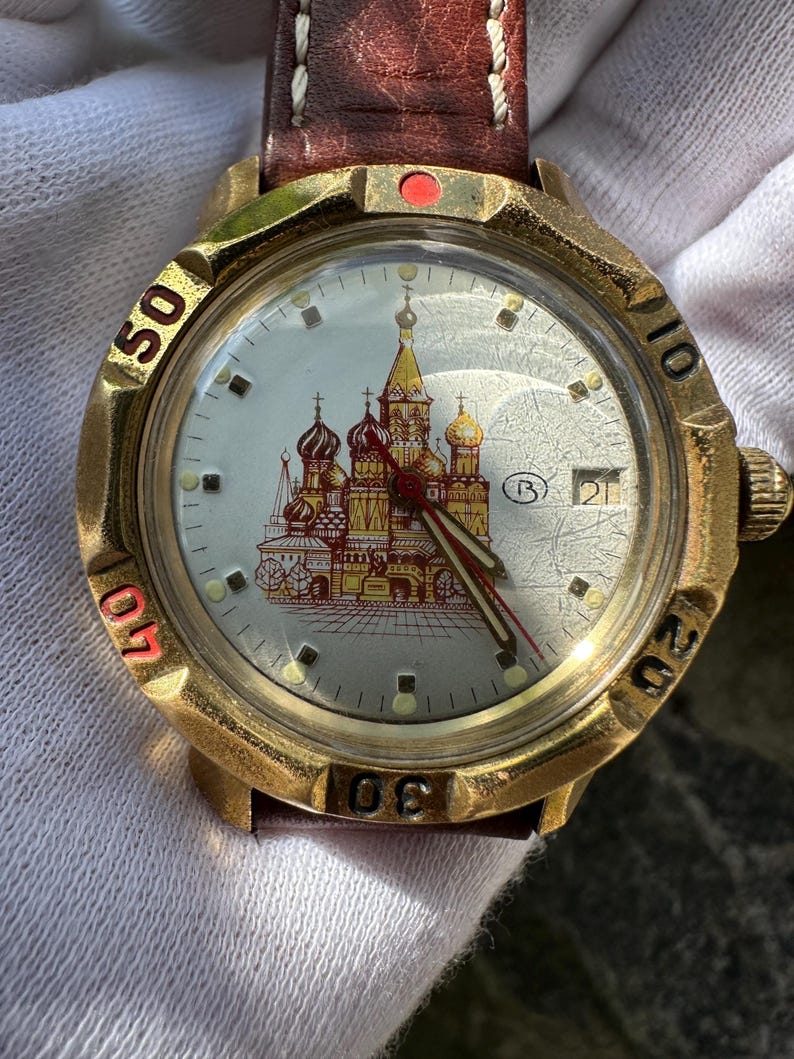 May include: A gold-toned wristwatch with a brown leather strap. The watch face features a detailed illustration of a cathedral with red and gold domes. The watch has a date display and red accents on the bezel.
