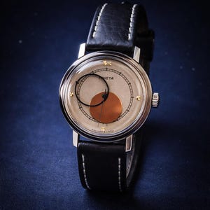 May include: A vintage wristwatch with a silver case and a black leather strap. The watch face features a light beige background with a copper-colored circle and black detailing. The watch has gold-colored accents and the word "PИETA" is visible.