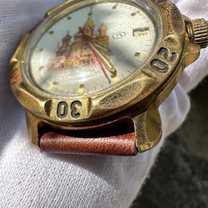 May include: Vintage gold-toned wristwatch with a brown leather strap. The watch face features a detailed illustration of a building with red and gold accents. The bezel has black numerals at the 20 and 30-minute marks.