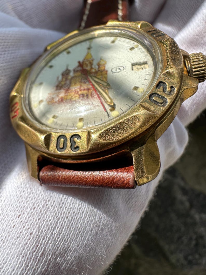 May include: Vintage gold-toned wristwatch with a brown leather strap. The watch face features a detailed illustration of a building with red and gold accents. The bezel has black numerals at the 20 and 30-minute marks.