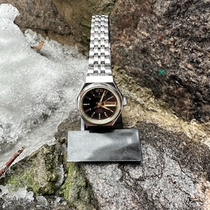 Könnte beinhalten: Eine silberfarbene Armbanduhr mit schwarzem Zifferblatt und Datumsanzeige. Die Uhr hat ein rechteckiges Zifferblatt und ein Metallarmband. Die Uhr wird auf einem schwarzen Ständer vor dem Hintergrund von Felsen und Schnee präsentiert.
