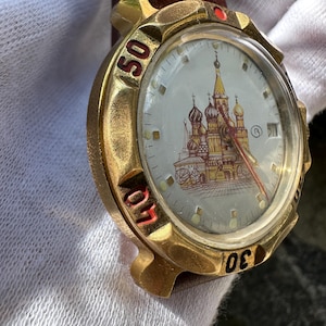 May include: A gold-toned wristwatch with a brown leather band. The watch face features a detailed illustration of a cathedral with red and gold accents. The numbers 30, 40, and 50 are visible on the bezel. The watch has a red second hand.