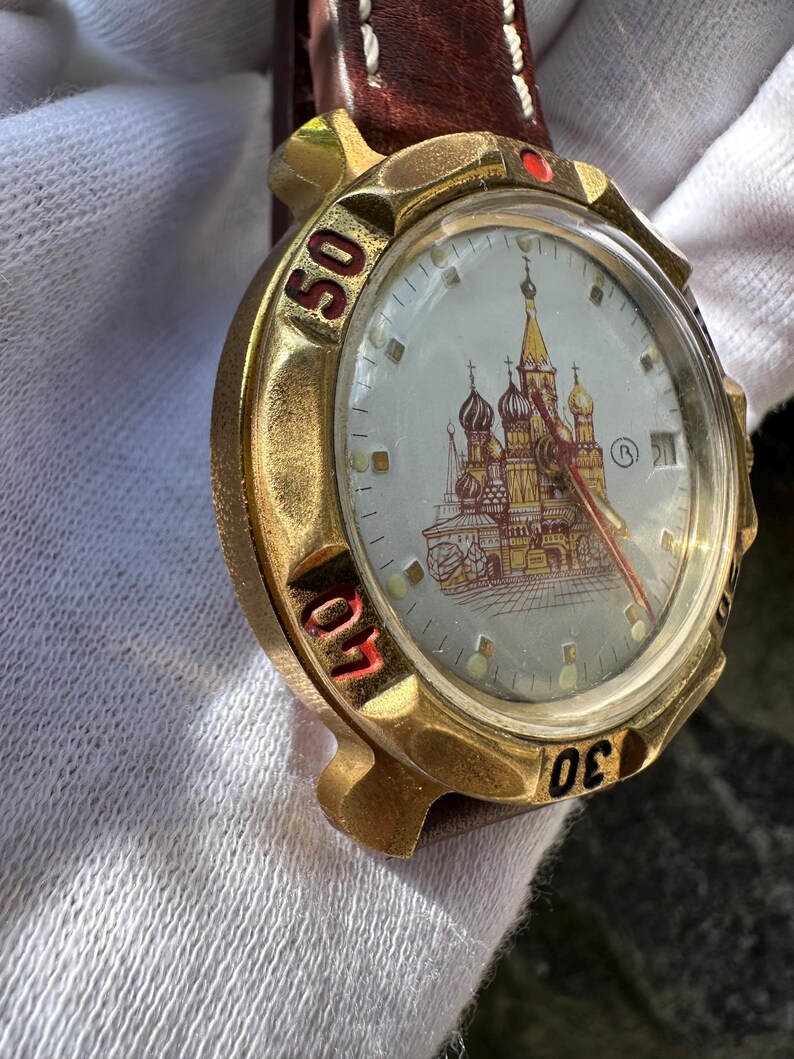 May include: A gold-toned wristwatch with a brown leather band. The watch face features a detailed illustration of a cathedral with red and gold accents. The numbers 30, 40, and 50 are visible on the bezel. The watch has a red second hand.