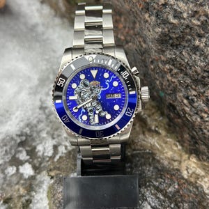 May include: A stainless steel wristwatch with a blue dial featuring an astronaut design. The watch has a blue bezel and a silver band. The date is set to Saturday, the 31st.