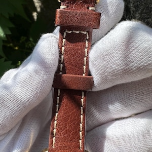 May include: Close-up of a brown leather watch strap with white stitching and a gold-colored buckle. The watch face is visible at the bottom, featuring a red accent and gold-colored casing. The strap is held by a gloved hand.