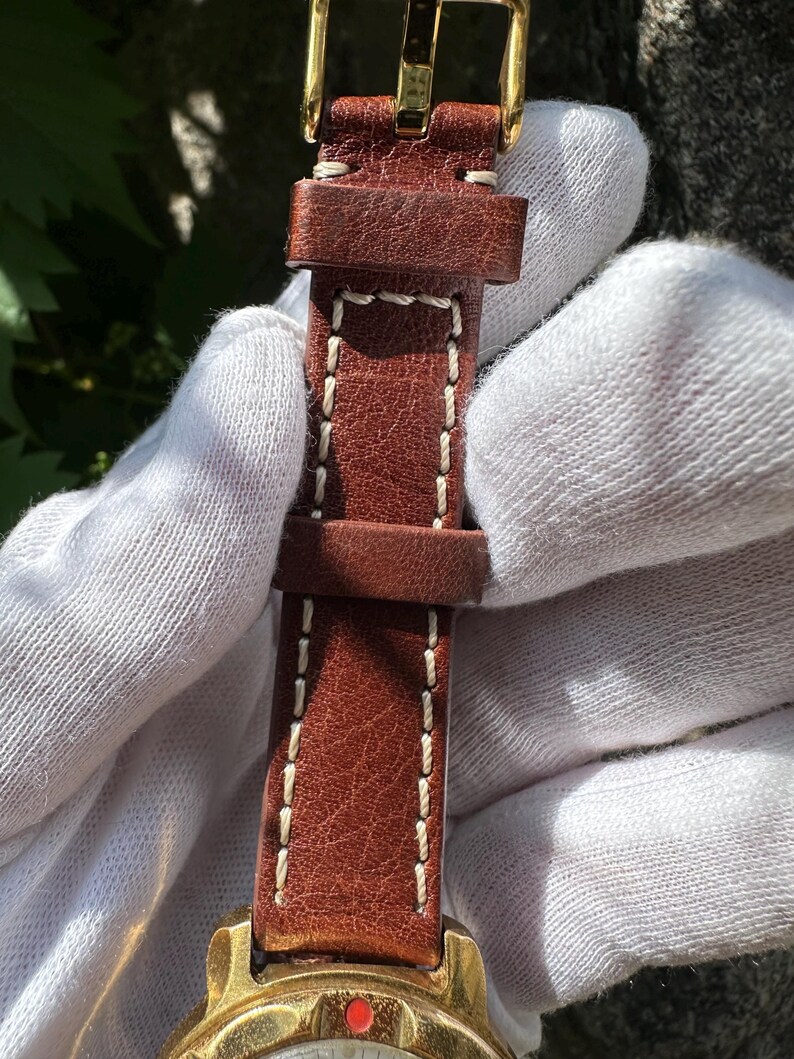 May include: Close-up of a brown leather watch strap with white stitching and a gold-colored buckle. The watch face is visible at the bottom, featuring a red accent and gold-colored casing. The strap is held by a gloved hand.