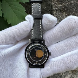 以下が含まれることがあります： 黒いラケタ腕時計、黒い革バンドと白いステッチ。文字盤は黒い背景に金と白のデザイン。時計にはシルバーのバックルが付いています。