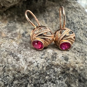 May include: A pair of rose gold earrings. Each earring features a filigree sphere with an oval, bright pink gemstone. The earrings are set against a textured gray stone background.