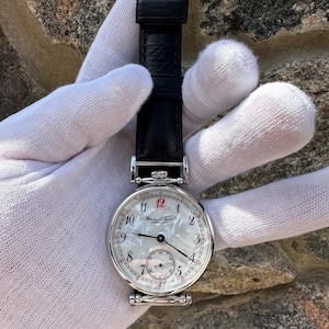Può includere: Un orologio da polso con cassa rotonda color argento, quadrante in madreperla bianca con numeri arabi neri, un numero 12 rosso e un piccolo quadrante dei secondi. L'orologio include un cinturino in pelle nera con fibbia color argento.