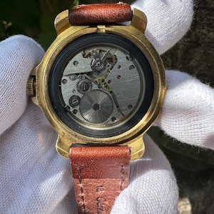 May include: Close-up of a vintage wristwatch with a gold-toned case and a brown leather strap. The watch face is open, revealing the intricate mechanical movement. The strap is approximately 0.75 inches wide.