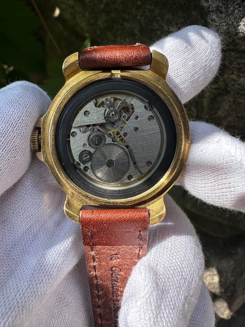May include: Close-up of a vintage wristwatch with a gold-toned case and a brown leather strap. The watch face is open, revealing the intricate mechanical movement. The strap is approximately 0.75 inches wide.