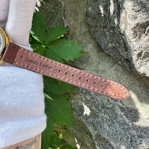 May include: Close-up of a brown leather watch strap with the text "18 Genuine Leather". The strap has several holes for adjustment and is stitched along the edges. The watch strap is brown.