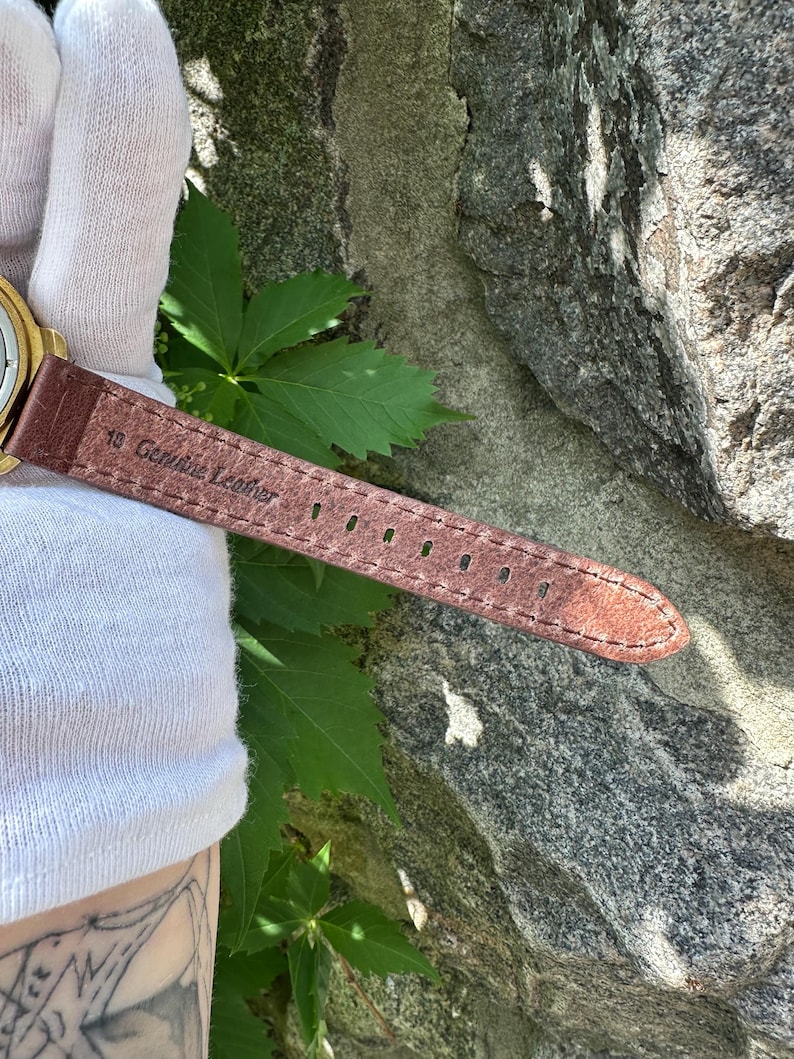 May include: Close-up of a brown leather watch strap with the text "18 Genuine Leather". The strap has several holes for adjustment and is stitched along the edges. The watch strap is brown.