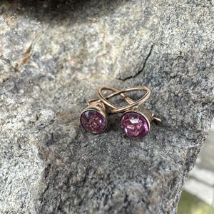 May include: A pair of gold-coloured stud earrings featuring round, faceted, pink gemstones. The earrings are displayed on a grey, textured rock surface. The gemstones have a deep pink hue, and the metal has a warm, polished finish.