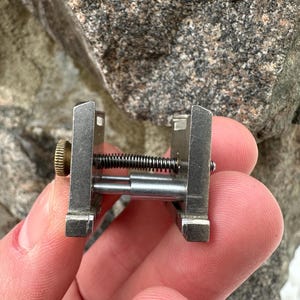 May include: A small, silver-coloured metal tool with a spring and a brass-coloured knob. The tool has a rectangular frame and a central spring mechanism. The object is held in a person's hand.