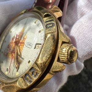 May include: Close-up of a vintage gold-toned wristwatch with a brown leather strap. The watch face features a detailed illustration. The bezel has black numerals. The watch is worn.