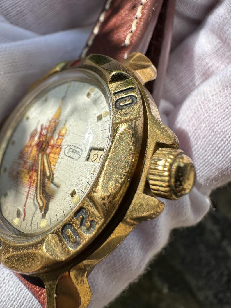 May include: Close-up of a vintage gold-toned wristwatch with a brown leather strap. The watch face features a detailed illustration. The bezel has black numerals. The watch is worn.