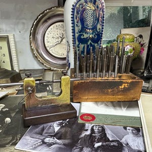 Puede incluir: Una variedad de objetos antiguos, incluyendo un reloj, un plato de plata, un jarrón decorativo con diseño de piña, un soporte de herramientas de madera con herramientas de metal y una cámara antigua. También se ven varias fotografías antiguas.