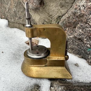 Può includere: Un utensile da orologiaio vintage in ottone con una parte superiore e una base color argento. L'utensile ha un braccio curvo e un quadrante. L'oggetto è appoggiato su una superficie innevata.