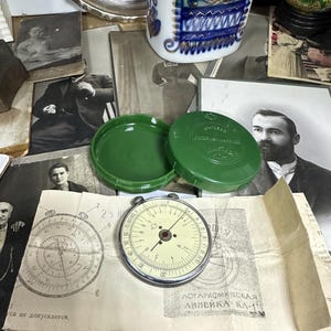 May include: A vintage slide rule with a cream dial and a silver frame, placed on an old document. Two green containers are open nearby. The background features antique black and white photographs and decorative objects.