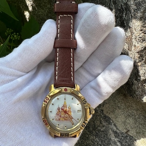 May include: A gold-toned wristwatch with a brown leather band and white stitching. The watch face features a detailed illustration of a building with red and gold accents. The watch band is approximately 8 inches long.
