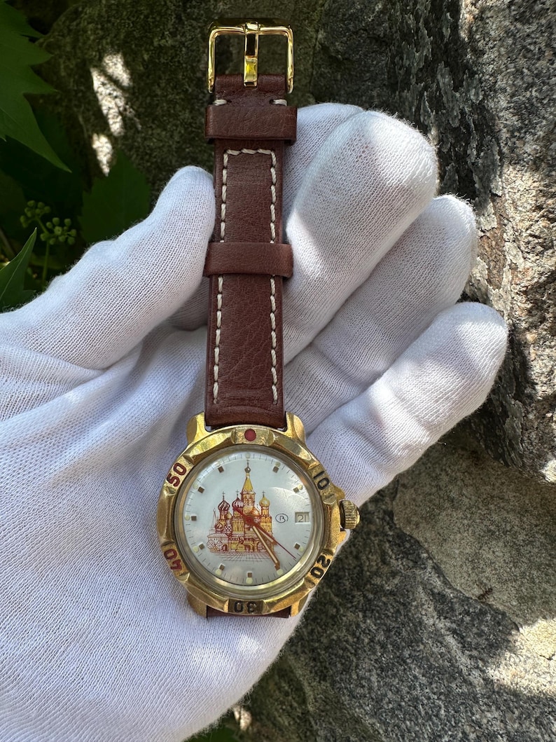 May include: A gold-toned wristwatch with a brown leather band and white stitching. The watch face features a detailed illustration of a building with red and gold accents. The watch band is approximately 8 inches long.