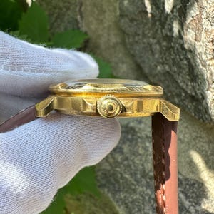 May include: Side view of a gold-toned wristwatch with a brown leather band. The watch has a textured face and a decorative crown. The watch is held by a gloved hand, showcasing the intricate details of the timepiece.